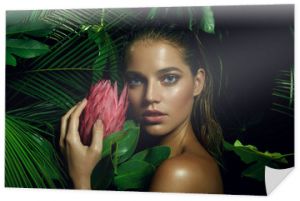 Piękna opalona dziewczyna z naturalnym makijażem i mokrymi włosami stoi w dżungli wśród egzotycznych roślin.