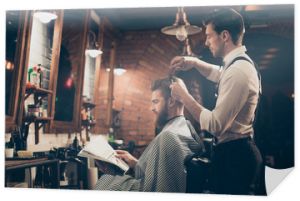 Niskokątny widok z profilu czerwonego brodatego klienta stylowego fryzjera, który otrzymuje idealną fryzurę od elegancko ubranego stylisty, czyta magazyn i czeka na wynik