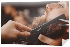 Hipster mężczyzna siedzi w fotelu fryzjera, podczas gdy fryzjer goli brodę nożyczkami.