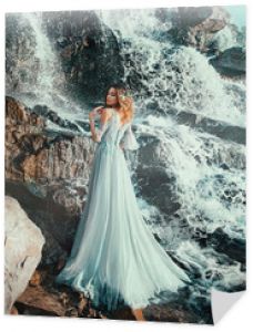 słodka dama w jasnoniebieskiej latającej sukience, nimfa wodna stoi w pobliżu dużego wodospadu w sprayu czystej zimnej wody. syrenka z ufarbowanymi na blond włosami stoi tyłem do aparatu i odwróconą twarzą