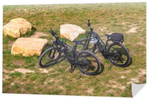 Dwa rowery elektryczne stoją na łące w pobliżu kamieni. Widok góry. Hełmy rowerowe wiszą na kierownicy.