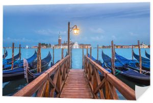 Wieczorny pejzaż morski Wenecji z gondolami na cumowaniach, światło latarni na drewnianym moście, widok na słynną wyspę San Giorgio Maggiore z bazyliką i dzwonnicą z Piazza San Marco. Włochy, Europa