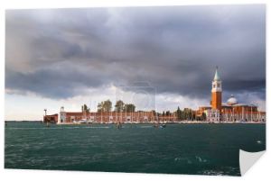 Wenecja, panorama wyspy San Giorgio Maggiore oglądane z wody, z łodzi. Campanile Bell Tower, kościół i latarnia morska Faro na wyspie San Giorgio Maggiore z błękitnym, burzliwym niebem w tle.