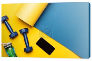 górny widok niebieskiej maty fitness z hantlami, smartfonem i butelką sportową na żółtym tle