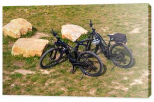 Dwa rowery elektryczne stoją na łące w pobliżu kamieni. Widok góry. Hełmy rowerowe wiszą na kierownicy.